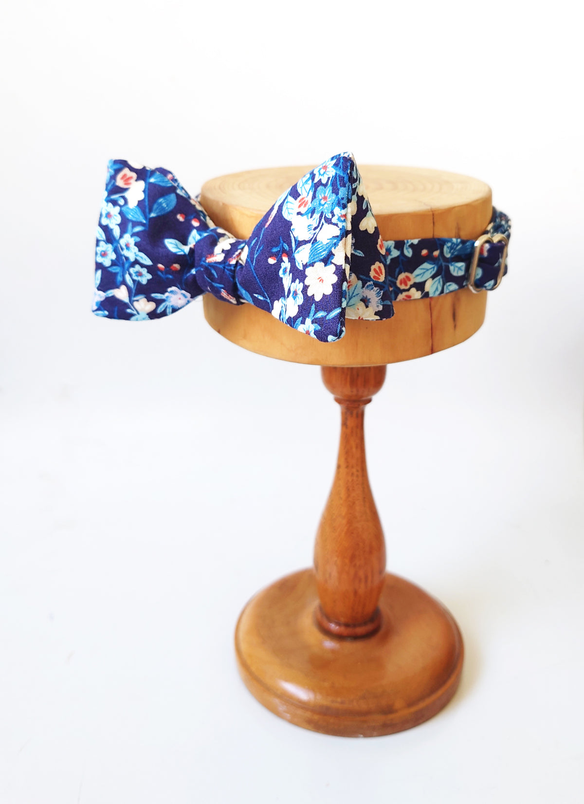 A navy blue bow tie with a white and light blue floral pattern, displayed on a wooden stand against a white background.