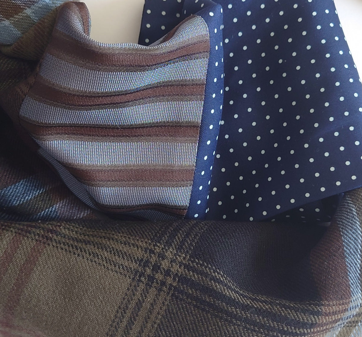 A folded woven scarf with multiple pattern sections: a wool plaid section in brown, beige, and navy, a striped section in taupe and brown silk, and a navy section with small white polka dots silk.