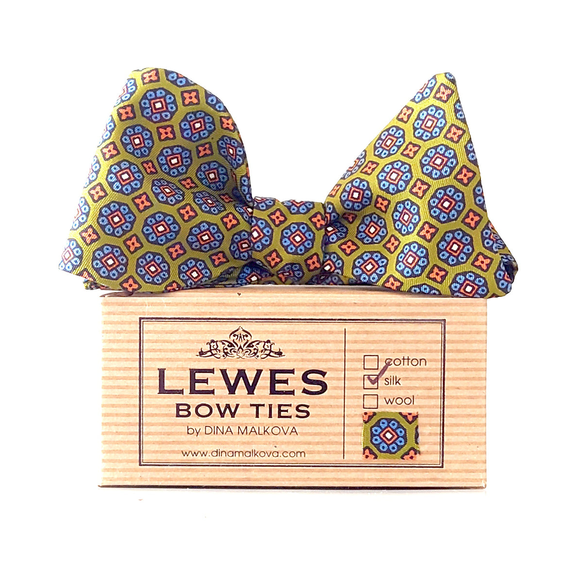 A patterned bow tie displayed on top of Lewes Bow ties brand box. The bow tie has an olive green base with a repeating geometric medallion pattern in blue, red, and orange.