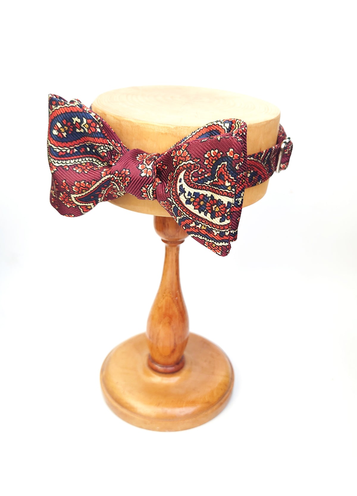 A paisley patterned bow tie in orange, navy, and rusty red, displayed on a round wooden stand against a white background.