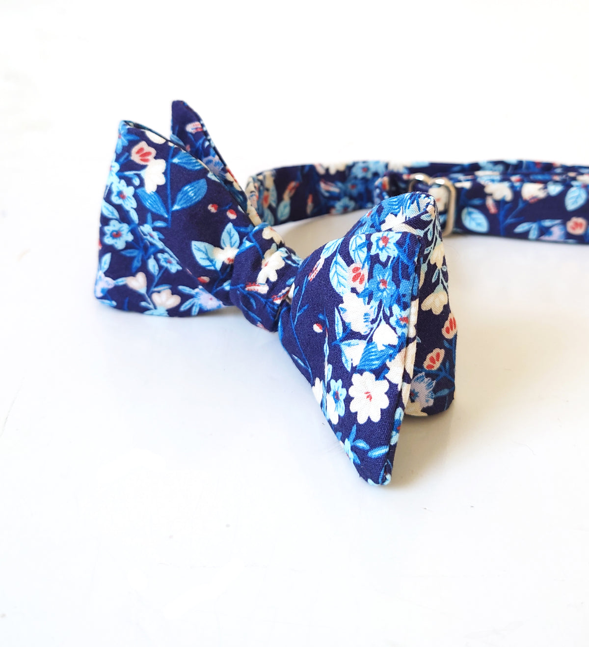A navy blue cotton bow tie with a white, light blue, and red floral pattern, displayed on a clean white background.